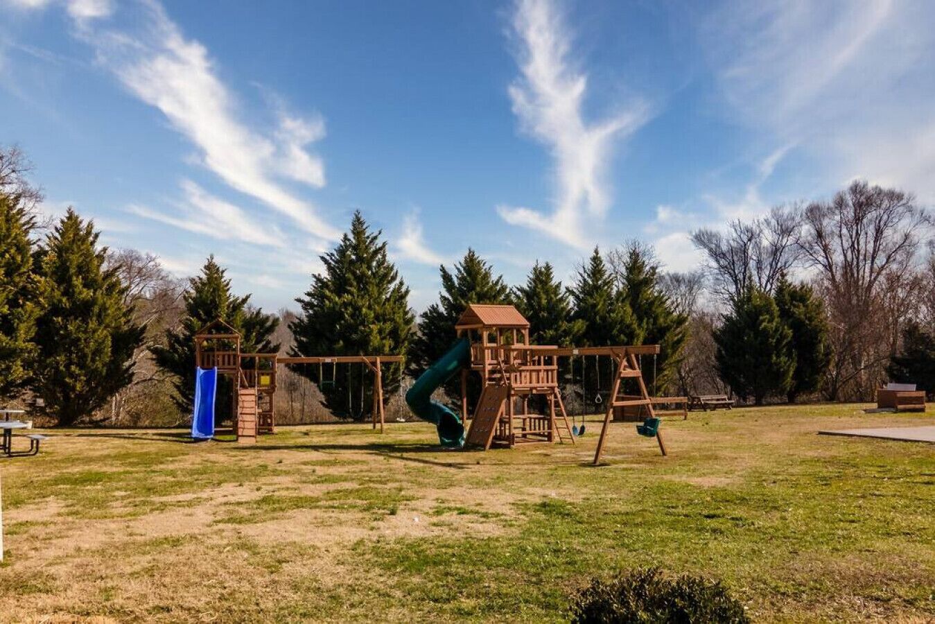Een speeltuin met een houten glijbaan en schommels, omringd door bomen en een picknicktafel.