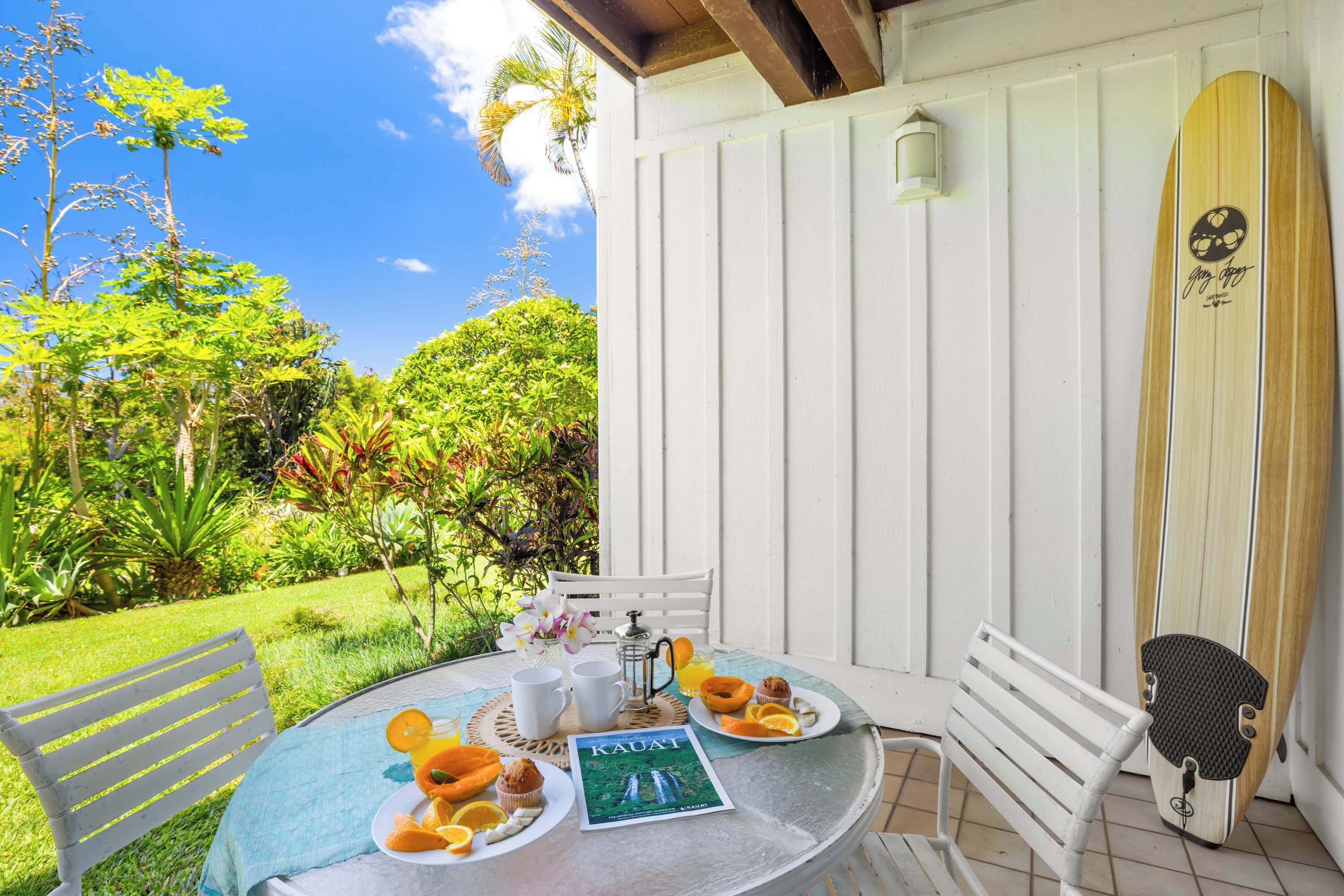 A table with breakfast items, a surfboard, and a magazine on a porch with a view of a garden and a white wall.