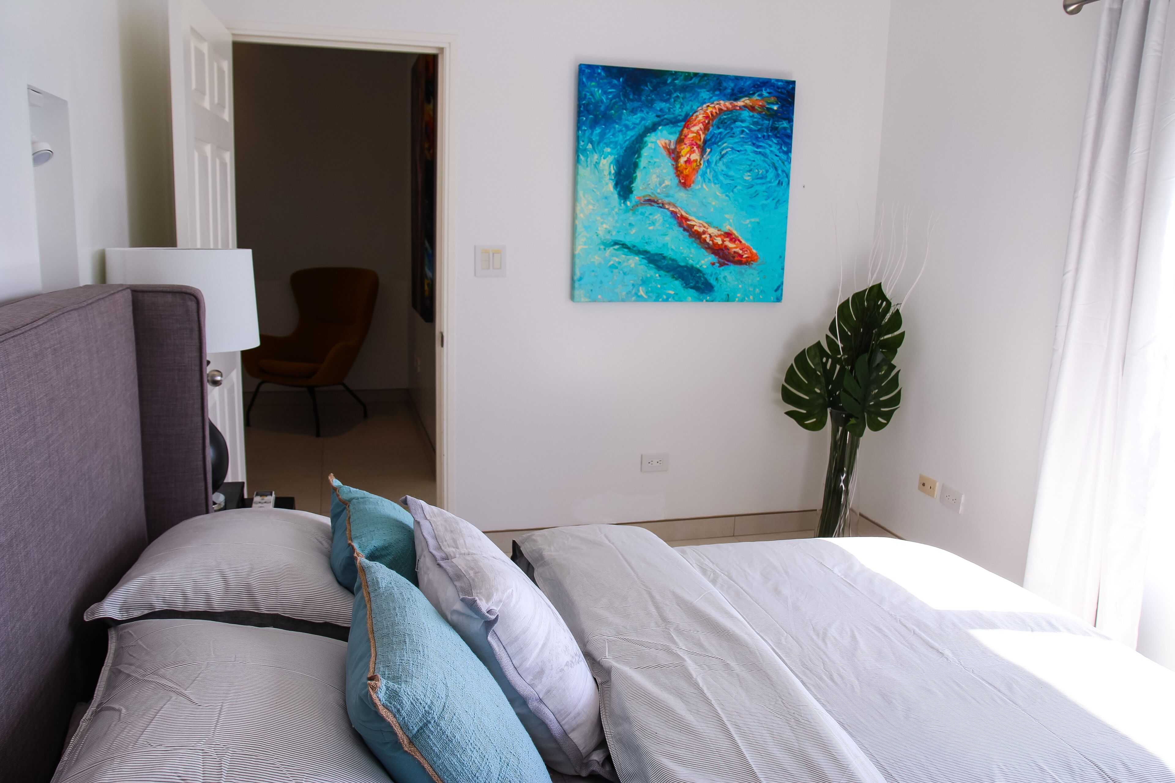 Una habitación de hotel con una cama, almohadas, un cuadro, una silla y una planta.