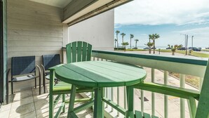 A balcony with a green table and chairs, a view of the beach, and a parking lot with cars.
