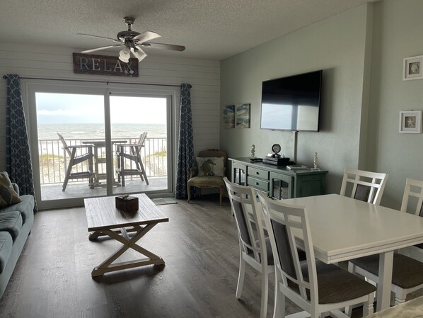 A room with a dining table, chairs, a ceiling fan, a TV, and a view of the ocean through a sliding door.