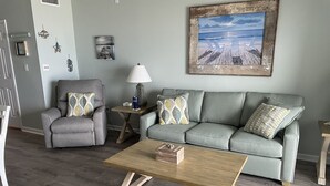 Un salón con un sofá gris, dos sillones, una mesa de centro de madera y un cuadro con una escena de playa.
