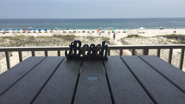 Una terraza de madera con un letrero de metal que dice "Beach" y vistas a la playa, con sombrillas y gente.