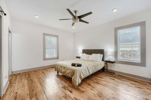 Un dormitorio con cama, ventilador de techo, dos lámparas de noche y ventanas amplias.