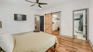 A bedroom with a bed, a ceiling fan, a TV, and a wooden door leading to a bathroom.