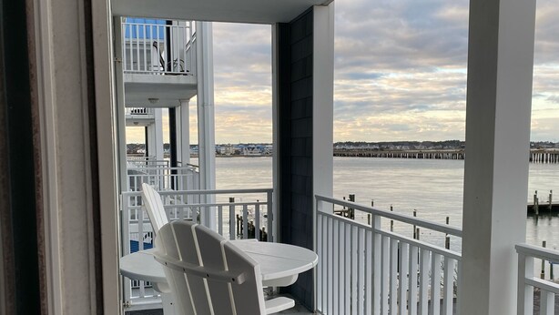 Een balkon met witte stoelen en een tafel, met uitzicht op een waterkant met een steiger.