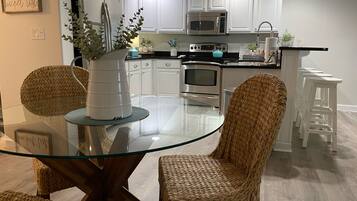 A modern kitchen with white cabinets, stainless steel appliances, and a glass dining table with wicker chairs.