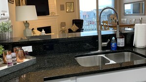 Een moderne keuken met een zwart granieten aanrecht, een roestvrijstalen spoelbak en witte kastjes. Door het raam is een eethoek met een tafel en stoelen zichtbaar.