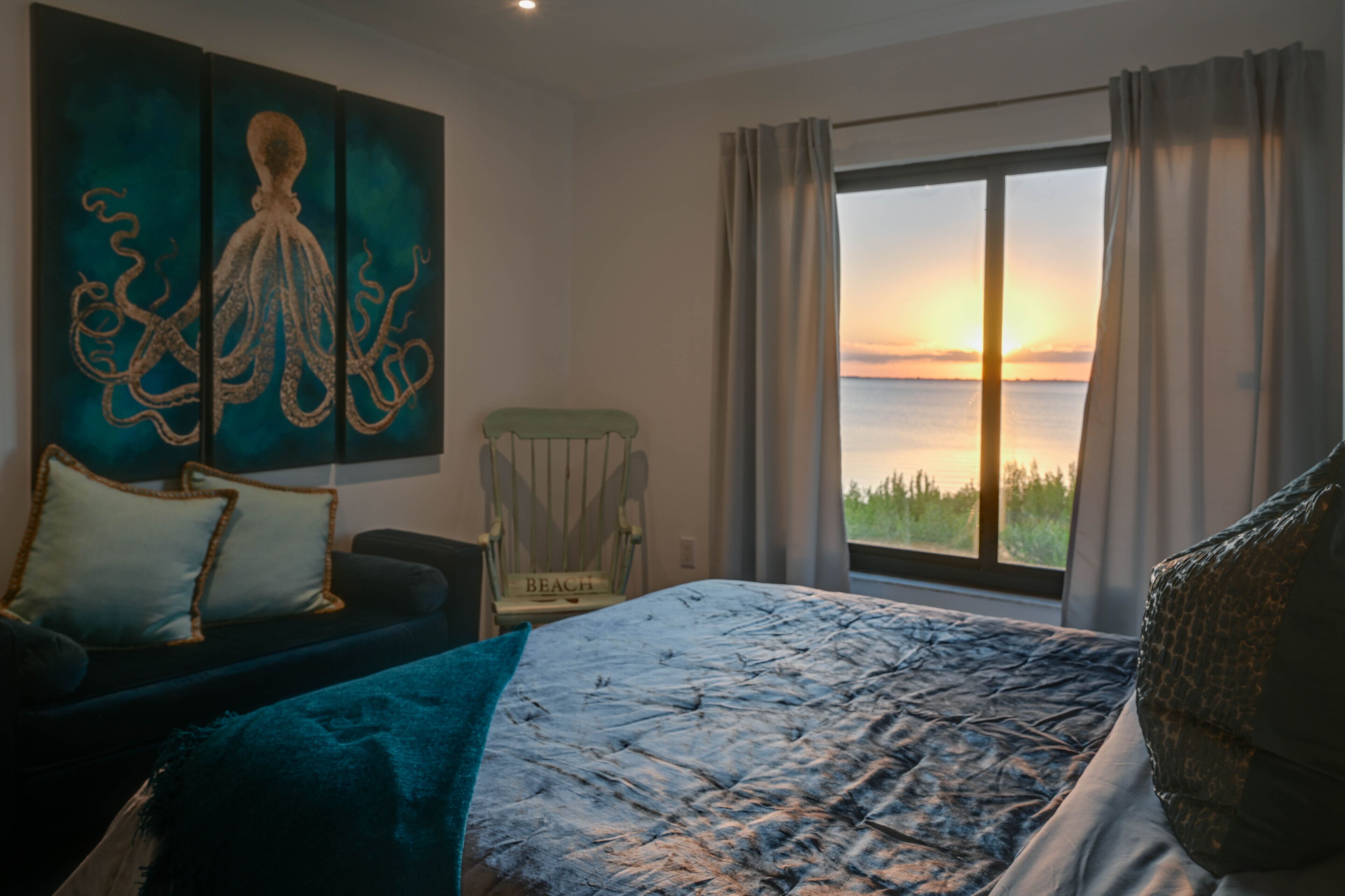Ein Schlafzimmer mit einem gro ßen Bett, einem Sessel, einem Gemälde und Blick auf den Sonnenuntergang.