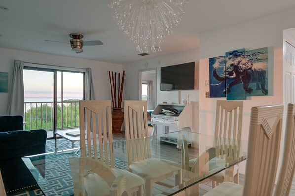A modern living room with a glass dining table, white chairs, a ceiling fan, a chandelier, and a large window with a view. - Chic Waterfront Condo in Cape Canaveral - 5 min from the beach! (Cape Canaveral)