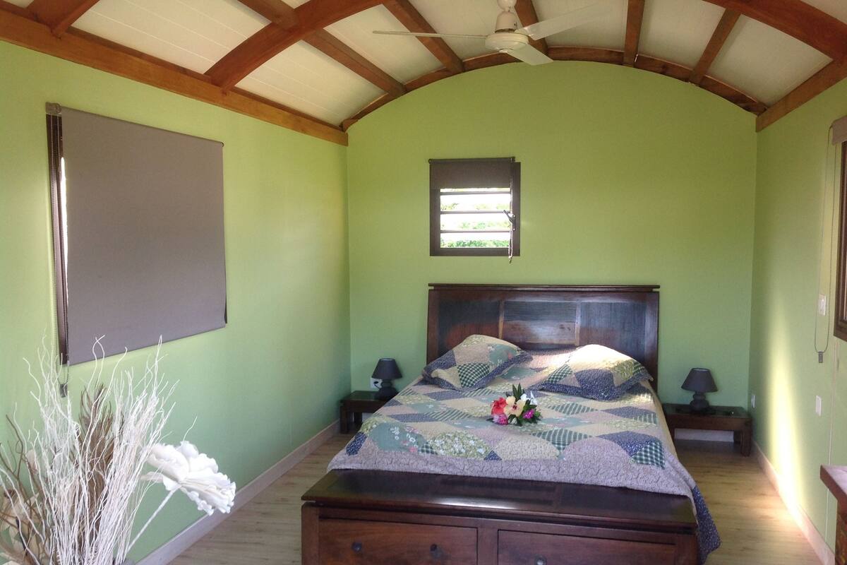 A bedroom with a wooden bed, a window, a ceiling fan, and a small table with a lamp.