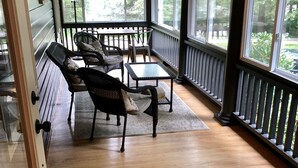 Une véranda couverte avec un plancher en bois, des meubles en osier et de grandes fenêtres donnant sur l’extérieur.