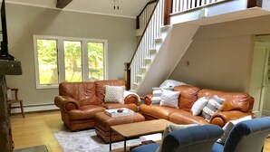 Un salon spacieux avec un canapé en cuir marron, des fauteuils bleus et une table basse en bois. Un escalier avec des rampes blanches est visible au fond.