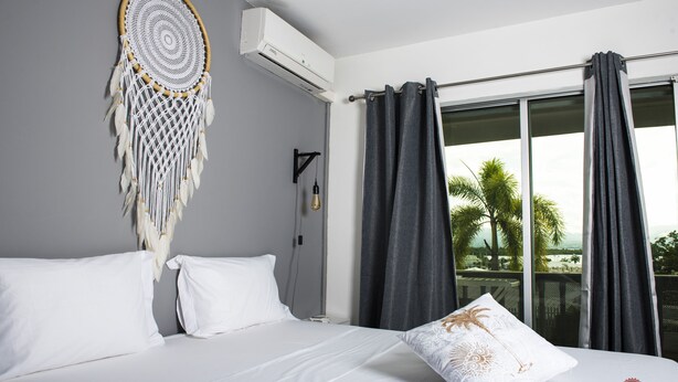 A hotel room with a bed, white bedding, a dreamcatcher, an air conditioner, a nightstand, a lamp, and a view of palm trees through the window.