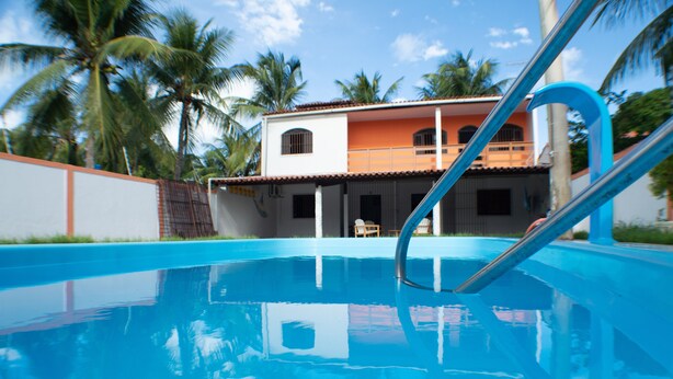 A swimming pool with a clear view of a two-story building in the background, surrounded by palm trees. - HOUSE ON FRENCH BEACH (Praia do Francês)