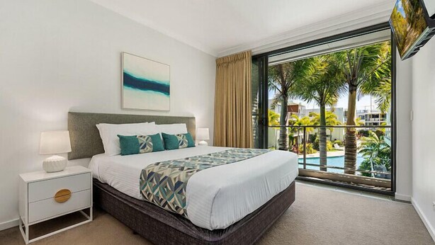 A hotel room with a large bed, a nightstand, a painting, and a view of a pool and palm trees. - 3 Bedroom Poolside Villa (Sunshine Coast)