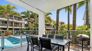 A balcony with a glass table and chairs, overlooking a swimming pool and palm trees. - 3 Bedroom Poolside Villa (Sunshine Coast)
