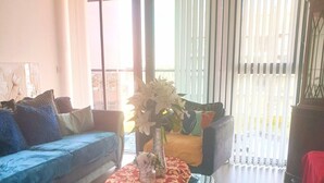 A living room with a sofa, a coffee table with a vase of flowers, and a view through a window with blinds. - New Modern Penthouse apartment with a fantastic view (Sutton)
