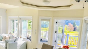 Ein Essbereich mit großem Tisch, Stühlen und Deckenventilator. Große Fenster bieten Blick auf einen Garten und eine Terrasse.