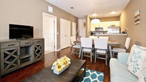 A living room with a TV, a dining area with chairs and a table, a kitchen with appliances, and a sofa. - Lovely Townhouse in a resort located less than 10 minutes from Disney complex. (Kissimmee)