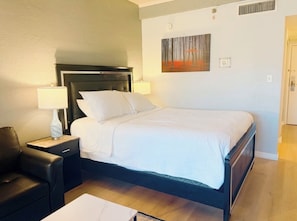 Quarto de hotel com cama, mesa de cabeceira com abajur, cadeira e quadro na parede.