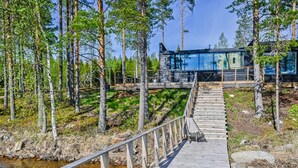 A wooden bridge leading to a modern cabin surrounded by tall trees and a clear stream.