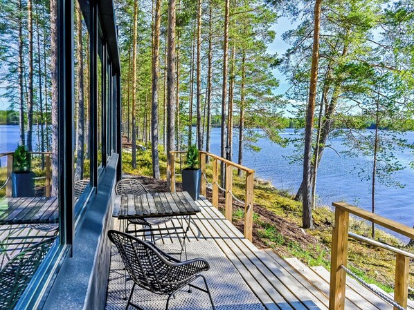 Una terraza con barandas de madera que da a un lago, amoblada con sillas y mesas de metal.