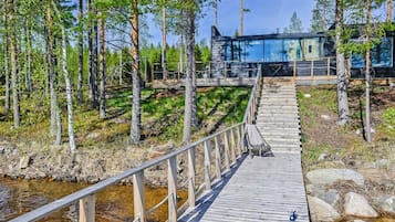 Una terraza de madera que conduce a una cabaña moderna con paredes de vidrio, rodeada de un bosque.