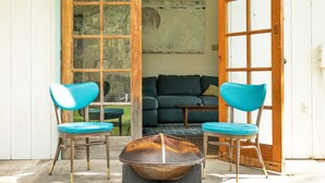 Een open deur die leidt naar een leefruimte met een bank, twee stoelen en een vuurkorf.