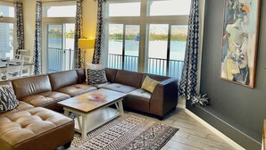 Un salon moderne avec un canapé en cuir marron, une table basse en verre et une vue sur un lac, visible à travers de grandes fenêtres.