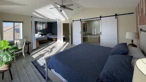 Une chambre à coucher avec un grand lit, un ventilateur de plafond, une fenêtre et une porte coulissante donnant sur une salle de bain.