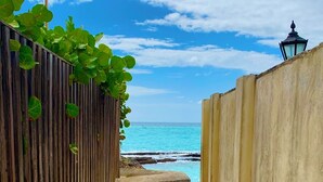 Uma trilha de areia estreita ladeada por cercas de madeira e um muro de concreto, que leva a uma praia com águas turquesa e céu claro.
