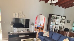 Uma sala de estar com um sofá azul, uma televisão, uma estante de livros e um espaço de jantar ao fundo.