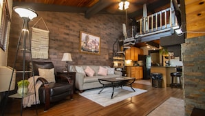A cozy living room with a leather sofa, a coffee table, and a brick wall. There is a staircase leading to an upper level.