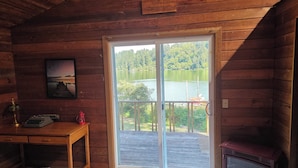 Una cabaña con interior de madera, un escritorio, una silla, una mesita, un calentador y vista a un lago a través de una puerta corrediza.