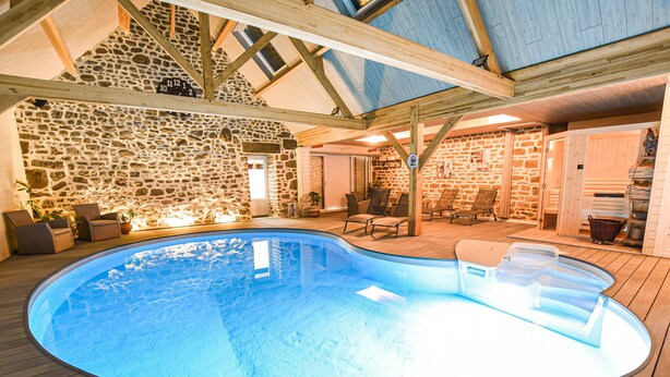 Una piscina interior con paredes de piedra y vigas de madera, rodeada de sillas de descanso y una pequeña zona de estar.