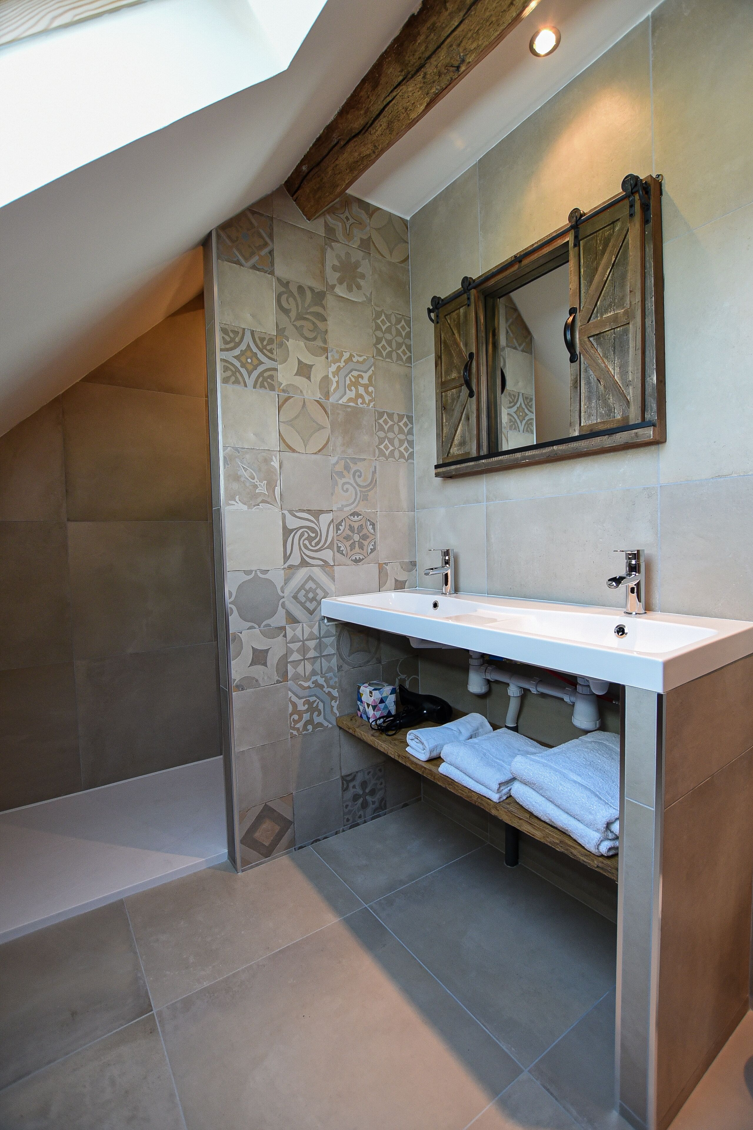 Une salle de bain moderne dotée d’une grande verrière, d’un plafond avec poutres apparentes, d’un meuble-lavabo à deux vasques et d’une étagère intégrée où sont rangées les serviettes.