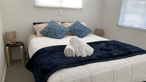 Een netjes opgemaakt bed met een donkerblauwe deken, witte kussens en een opgerolde witte handdoek. Er zijn twee nachtkastjes met lampen en een raam met jaloezieën.