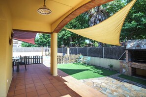 Un patio couvert avec une table et des chaises, un barbecue et un petit jardin agrémenté de gazon synthétique et d’un mur en pierre.