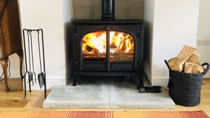 A wood-burning stove with a fire burning inside, a metal rack with tools, and a basket of firewood. - 5 Star Coachmans cottage. Built in the mid 18th Century. (Stranocum)