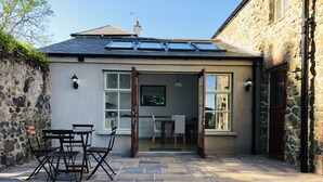 A stone building with a tiled patio, a dining area with a table and chairs, and a kitchen visible through an open door. - 5 Star Coachmans cottage. Built in the mid 18th Century. (Stranocum)