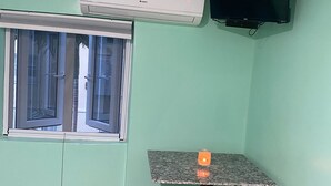 Una stanza piccola con una finestra, un tavolo, due sgabelli, un condizionatore d'aria e una TV fissata al muro.