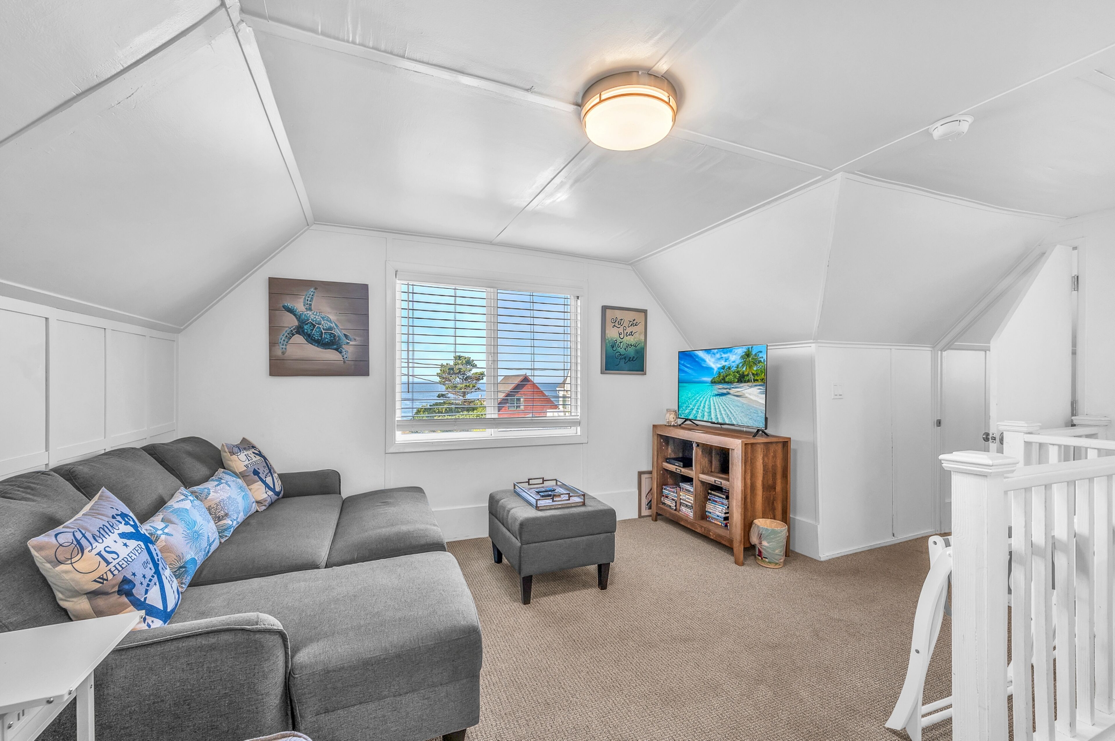 A cozy attic bedroom with a gray sectional sofa, a small ottoman, a wooden TV stand, and a white crib.