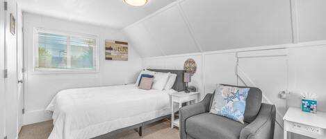 A bedroom with a bed, a gray armchair, a nightstand, and a window with blinds.