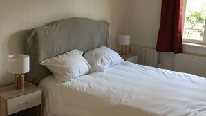 A neatly made bed with white linens, a wooden nightstand with a lamp, and a window with red curtains. - Domain House - Landin Castle (LE LANDIN)