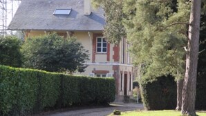 A house with a sloped roof, a small window, and a door. There is a hedge in front of the house and a tree to the right. - Domain House - Landin Castle (LE LANDIN)