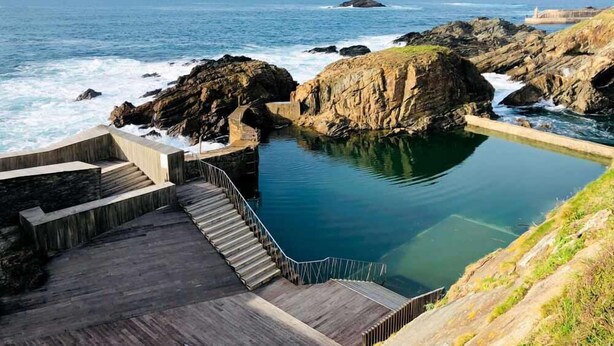 A coastal pool area with steps leading down to the water, surrounded by rocky terrain and overlooking the ocean.