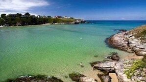 A coastal area with clear turquoise water, rocky shoreline, and houses on a hill.