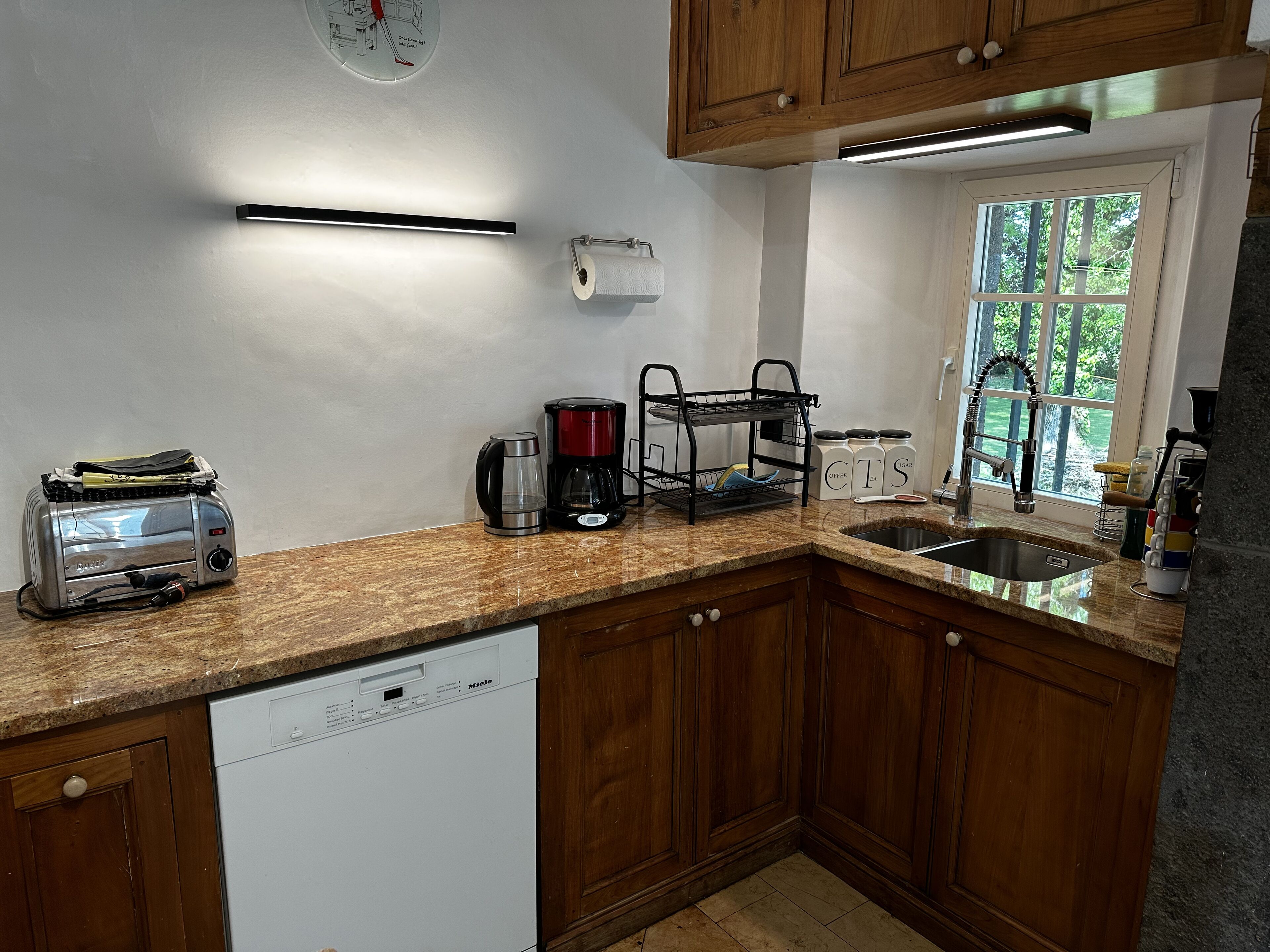 Una cocina con armarios de madera, lavavajillas integrado, cafetera, tostadora y ventana con vistas a árboles.