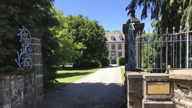 Un chemin de gravier mène à une entrée fermée par un portail en métal et un mur de pierre orné d’une plaque décorative en métal.